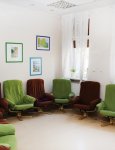 Wnętrze domu dziennego. Pokój wypoczynkowy dla klientów domu. Pod ścianami rzędy foteli wypoczynkowych w kolorach zielonym i brązowym. Na ścianach obrazy w kolorowych ramach.